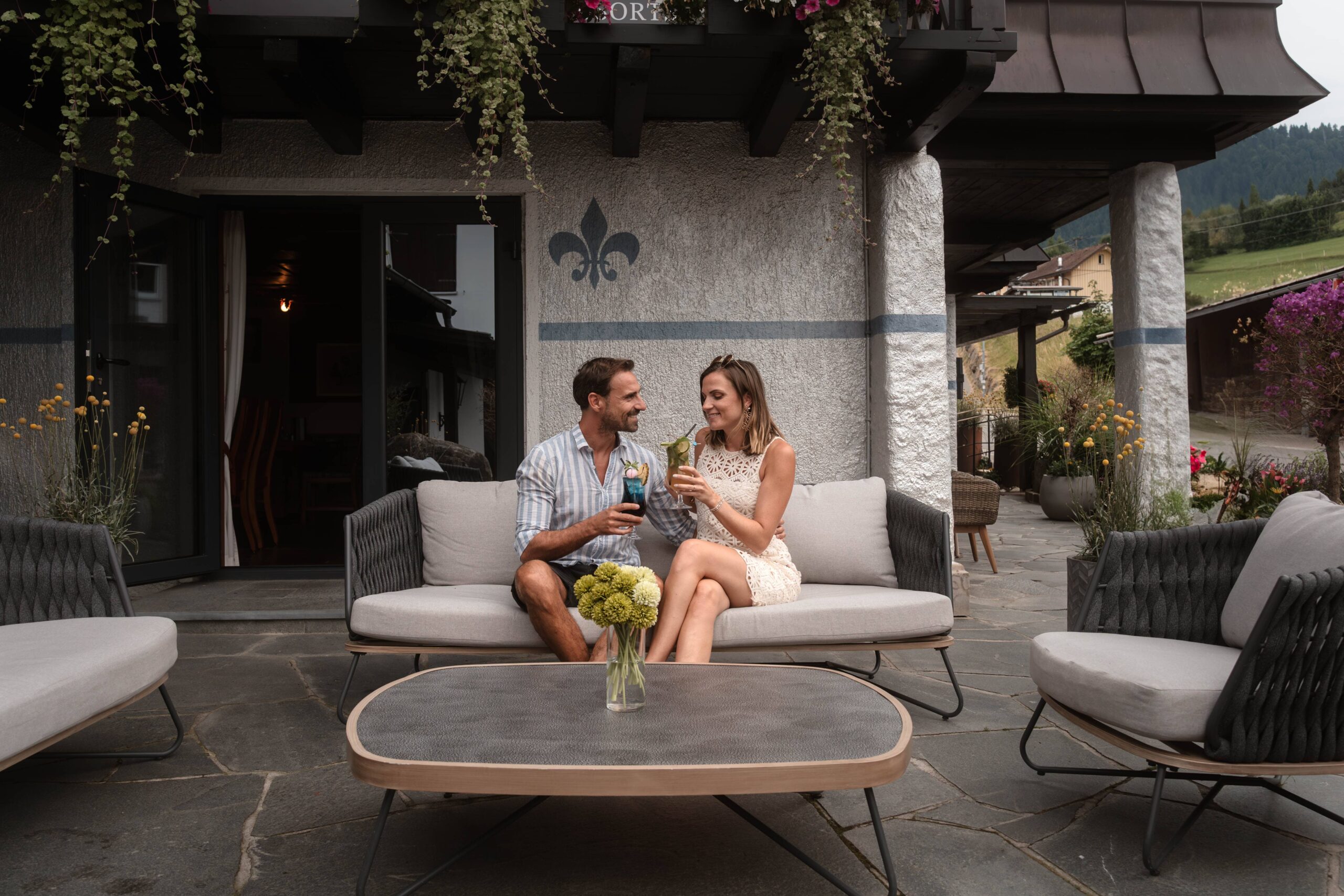 Paar genießt Cocktails auf der Terrasse des Hotel Alpenglück in den Alpen, umgeben von gemütlichen Möbeln und Blumen.