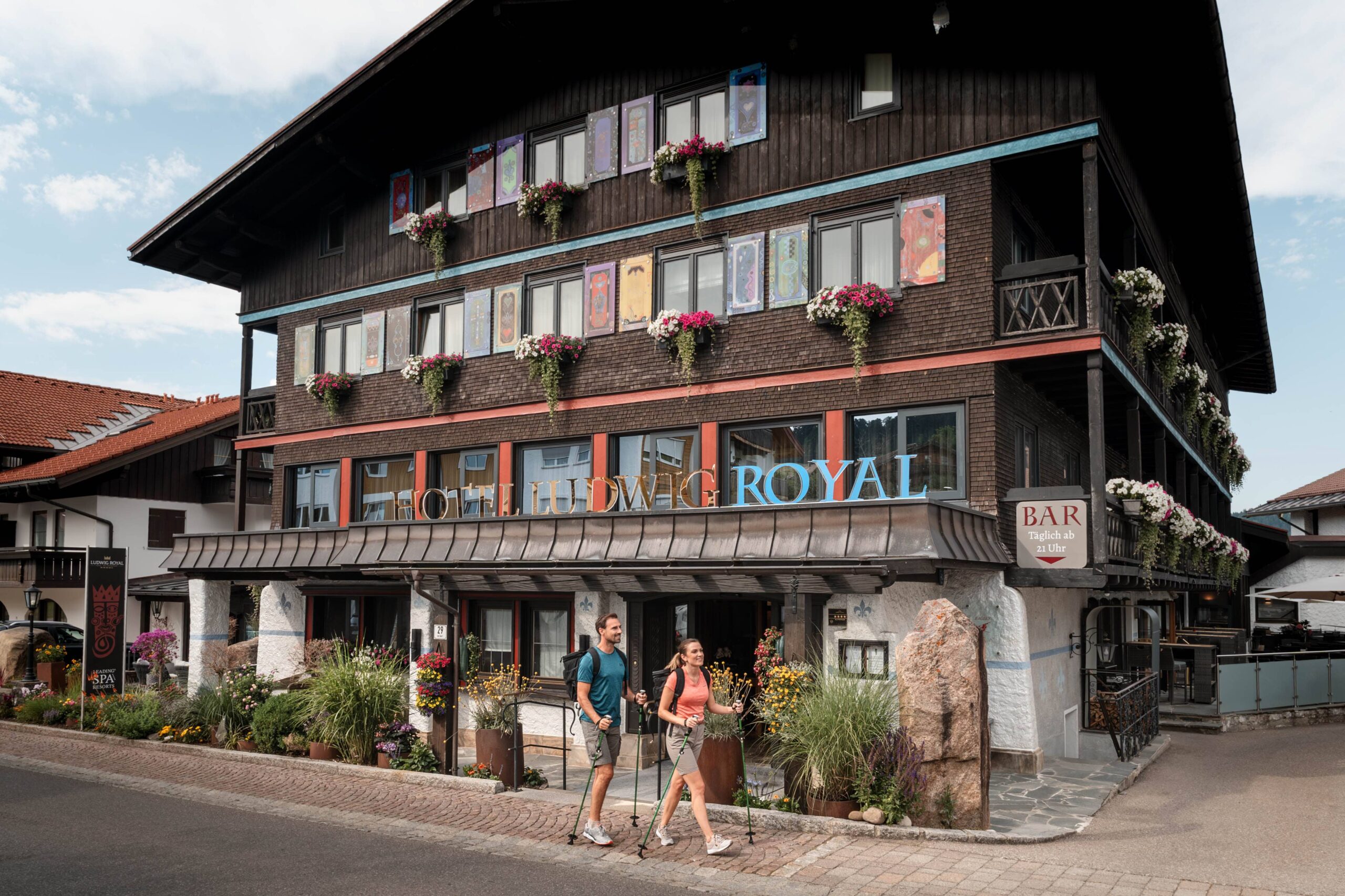Paar beim Wandern vor dem Hotel Ludwig Royal in Bayern, umgeben von Blumen, ideal für einen aktiven Familienurlaub.