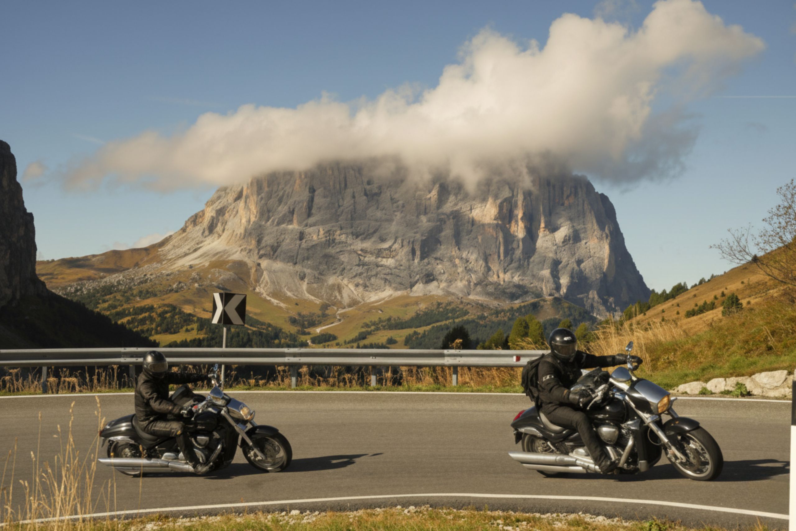 Zwei Motorradfahrer fahren auf einer kurvenreichen Straße in den Dolomiten, umgeben von majestätischen Bergen und schöner Natur.