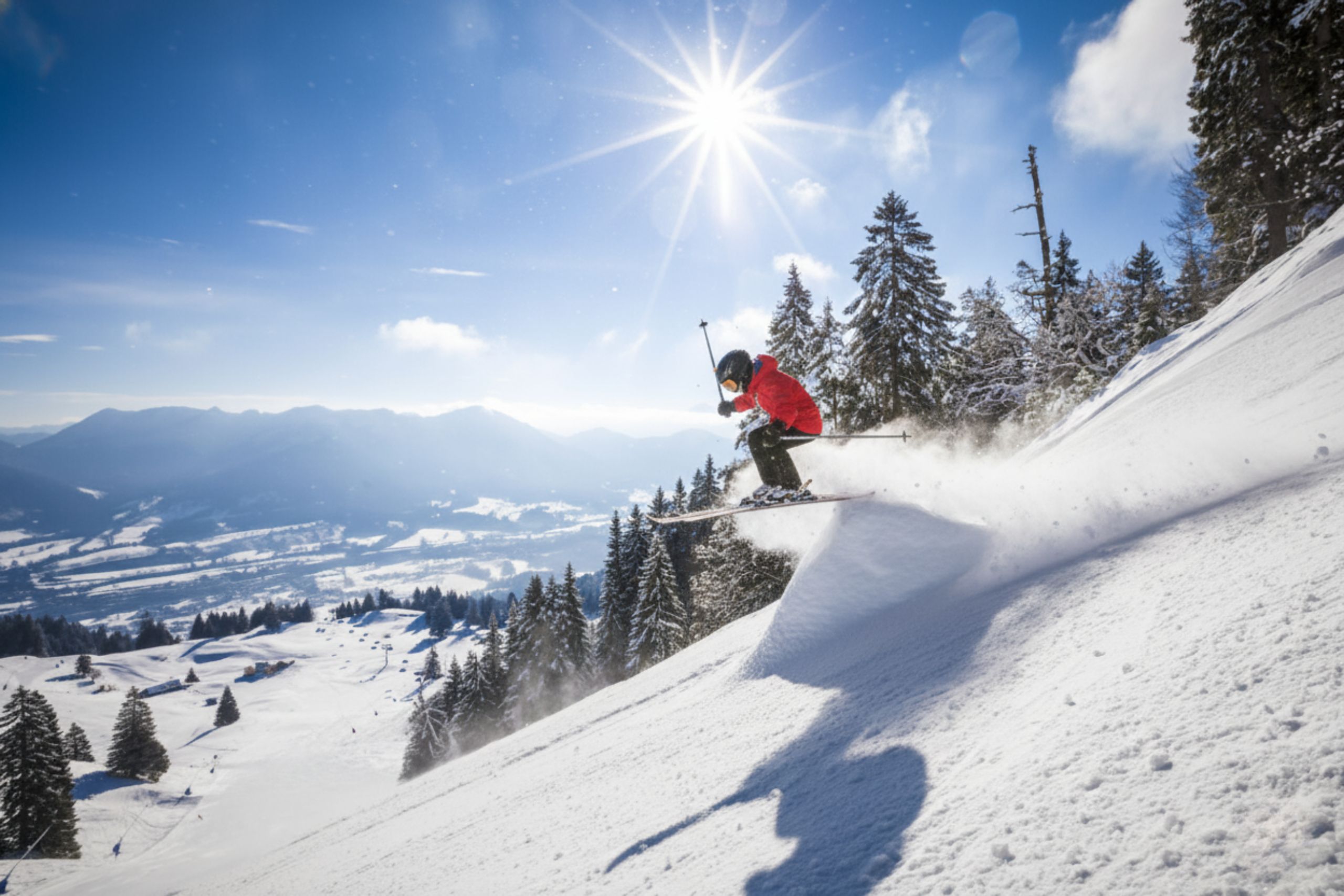 Ein Kind in roter Skijacke springt über eine Snowboardkante im verschneiten Alpengebiet, ideal für einen Winterurlaub.