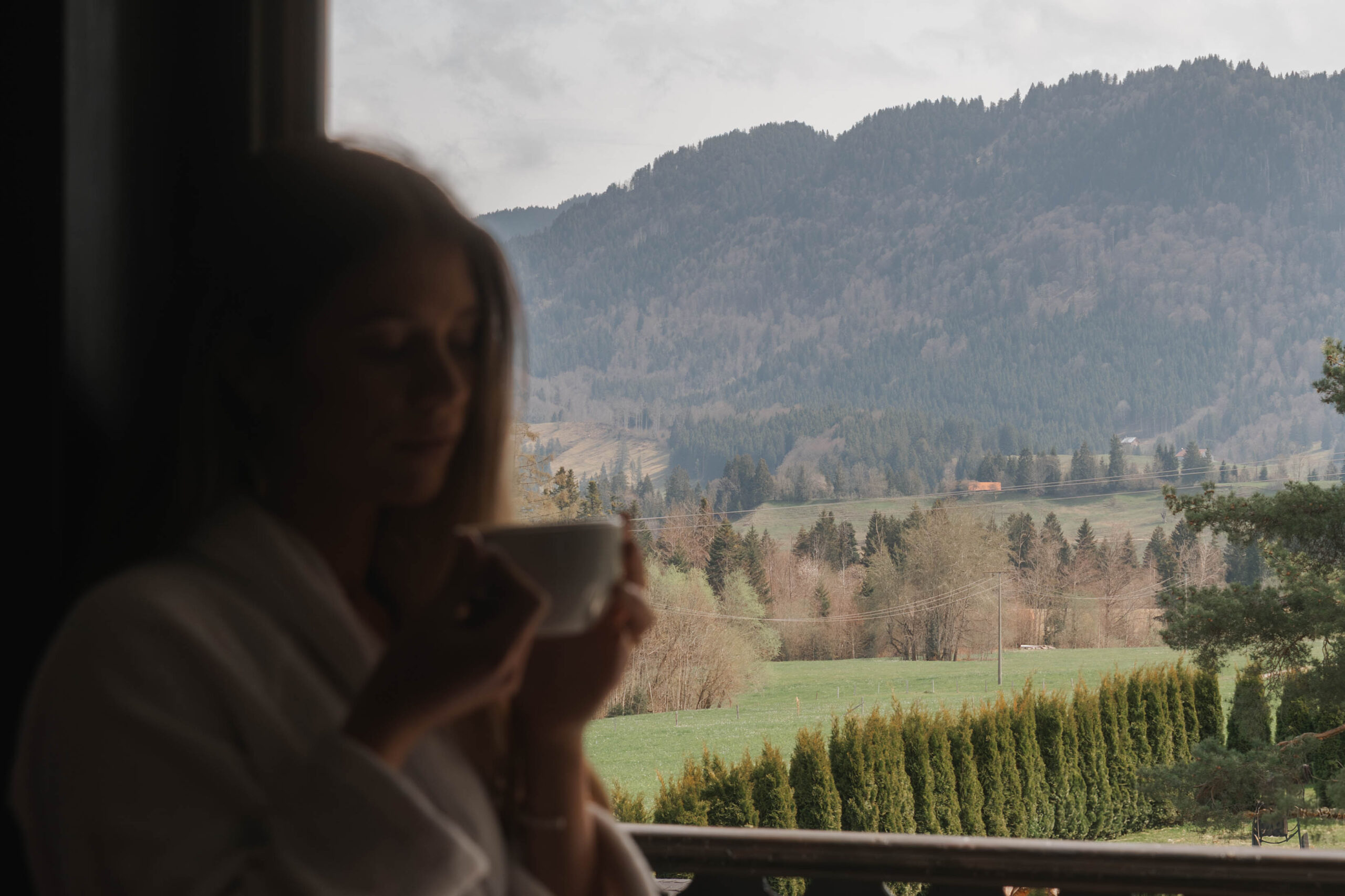 Frau mit einer Tasse in einem Hotelzimmer mit Blick auf die Berge und die malerische Natur im Familienurlaub.