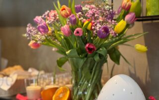 Bunte Tulpen in einer Vase im Hotel, umgeben von frischen Früchten und einem dekorativen Ei für den Familienurlaub.