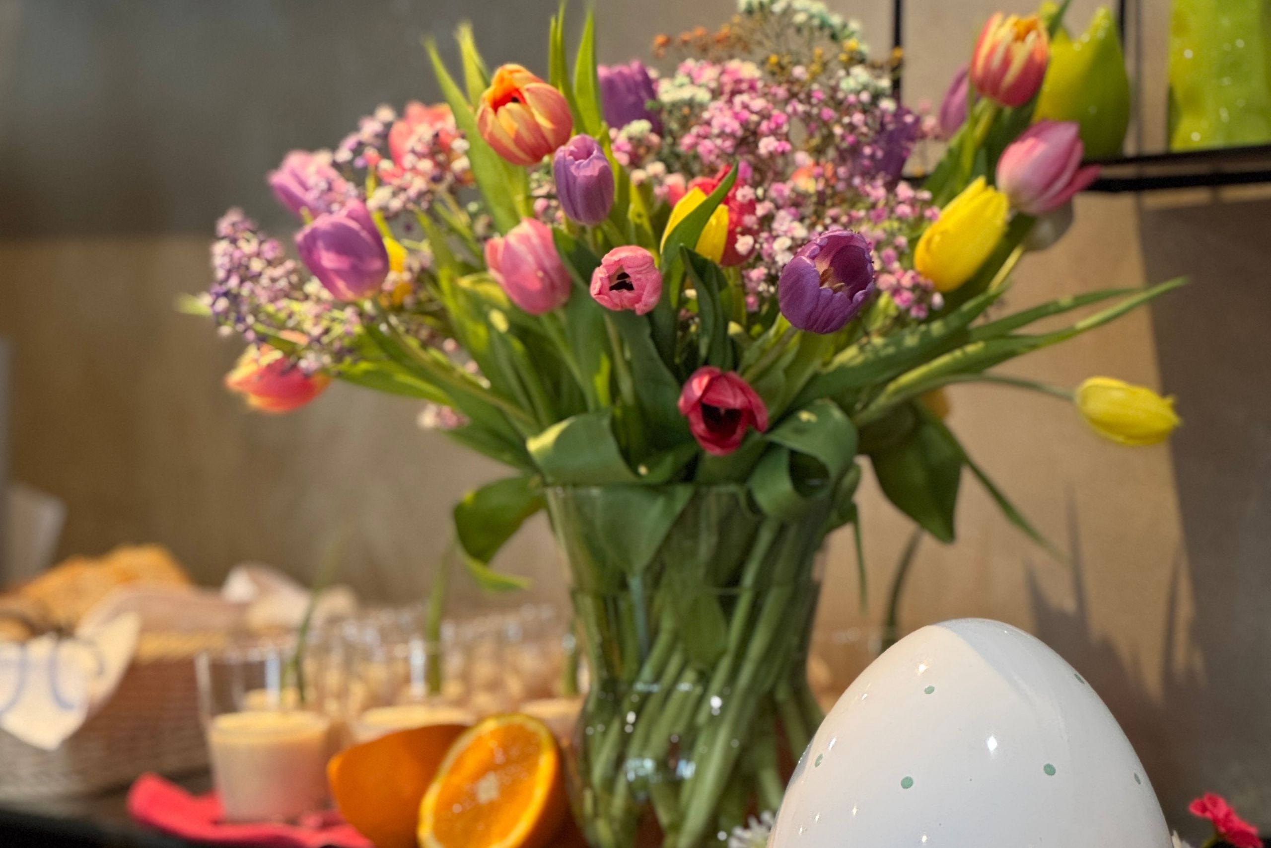 Bunte Tulpen in einer Vase im Hotel, umgeben von frischen Früchten und einem dekorativen Ei für den Familienurlaub.