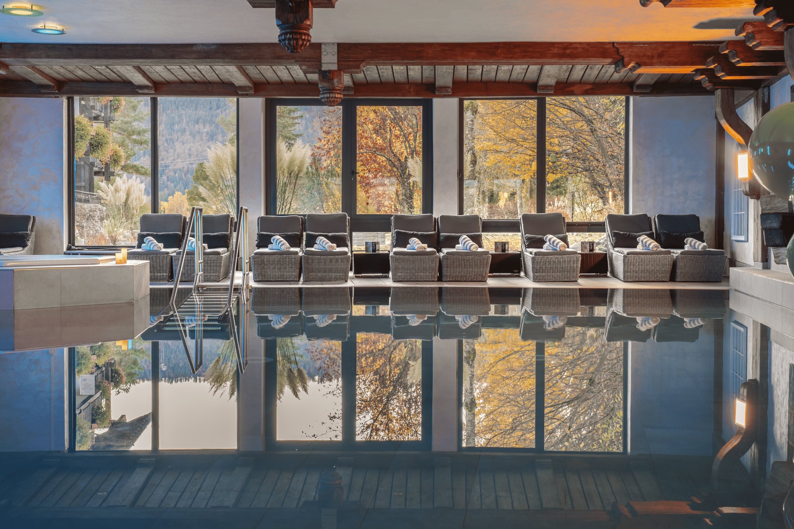 Wellnessbereich des Hotels mit Indoor-Pool und Liegen, umgeben von großen Fenstern und herbstlicher Natur im Hintergrund.